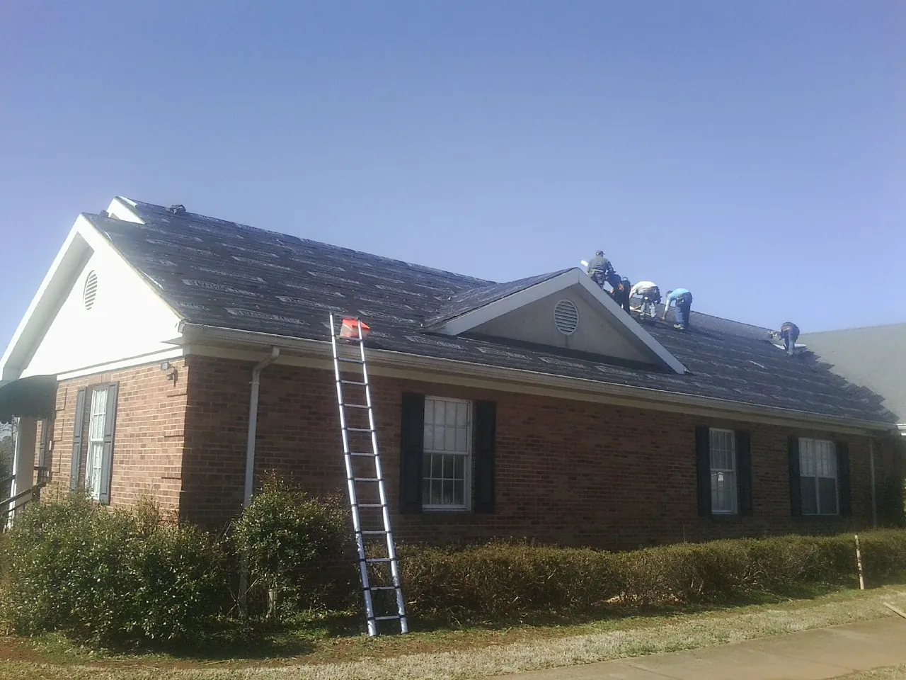 Crew tearing shingles off a brick ranch roof with a ladder set — Emergency Roof Repair work by our crew in Swink