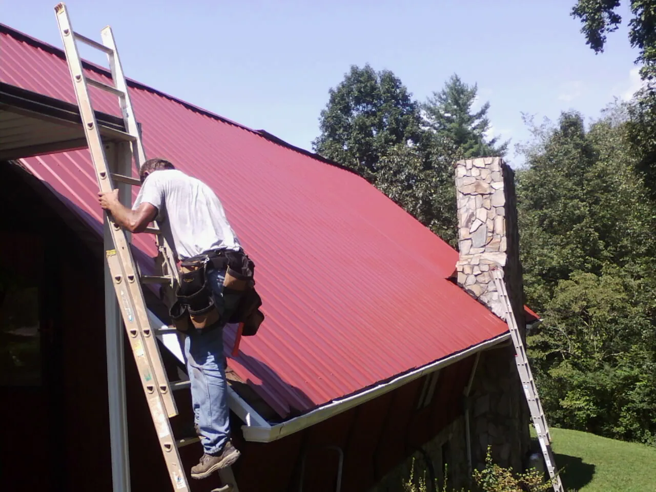 Roofer on a ladder beside a red metal roof and stone chimney — Soffit & Fascia Repair by Roofing Brothers in Swink